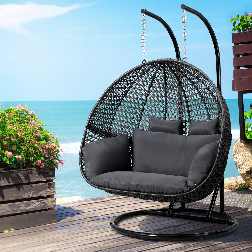 Outdoor Swing Chair Hanging Rattan Patio Furniture Pod Stand Cushion 2 Seater with black cushions on wooden deck by the sea