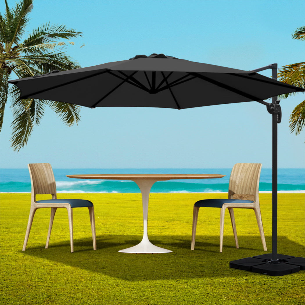 3m Outdoor Umbrella w/Base Cantilever Beach Roma 360 Degree Tilt Black with wooden chairs and white round table on grass by the sea