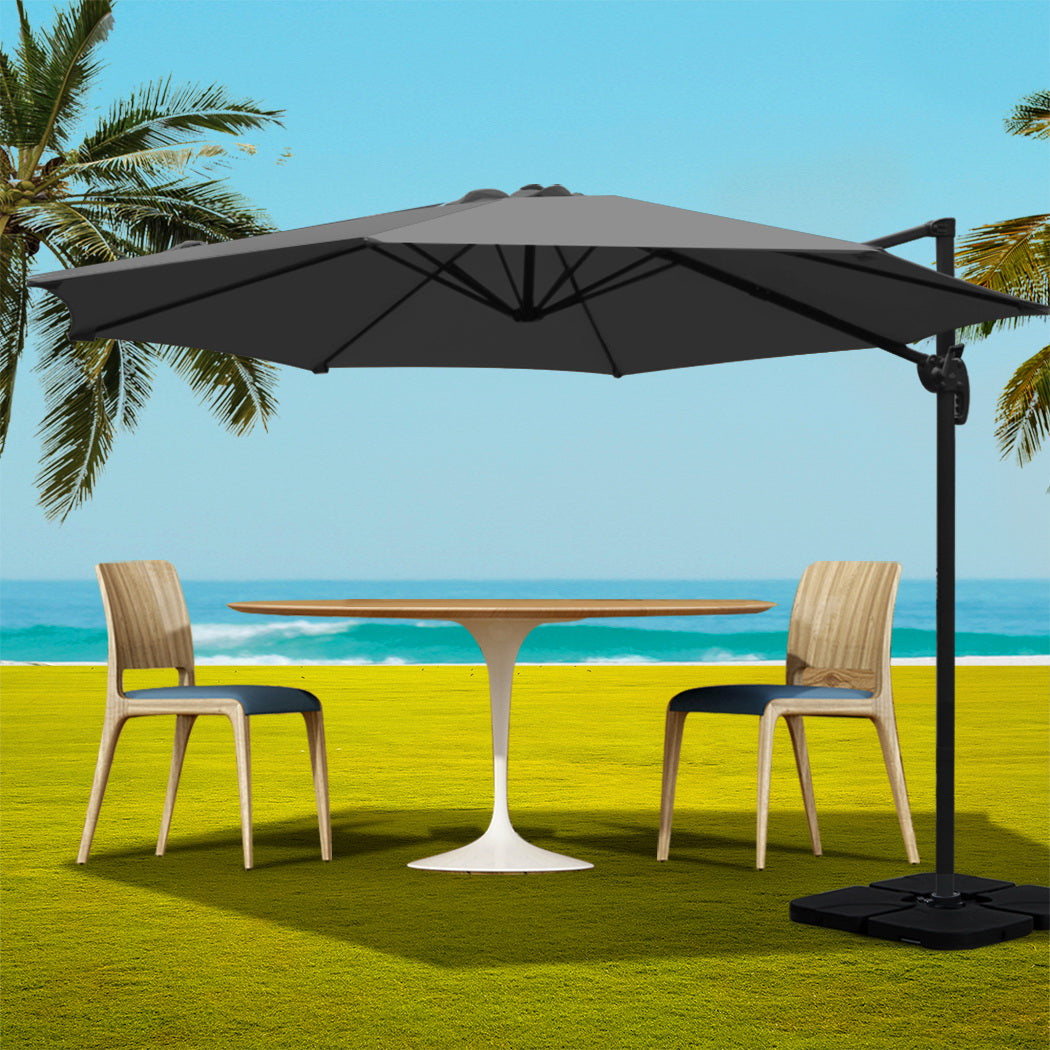 3m Outdoor Umbrella w/Base Cantilever Beach Roma 360 Degree Tilt Charcoal over patio table and chairs on grass by the sea