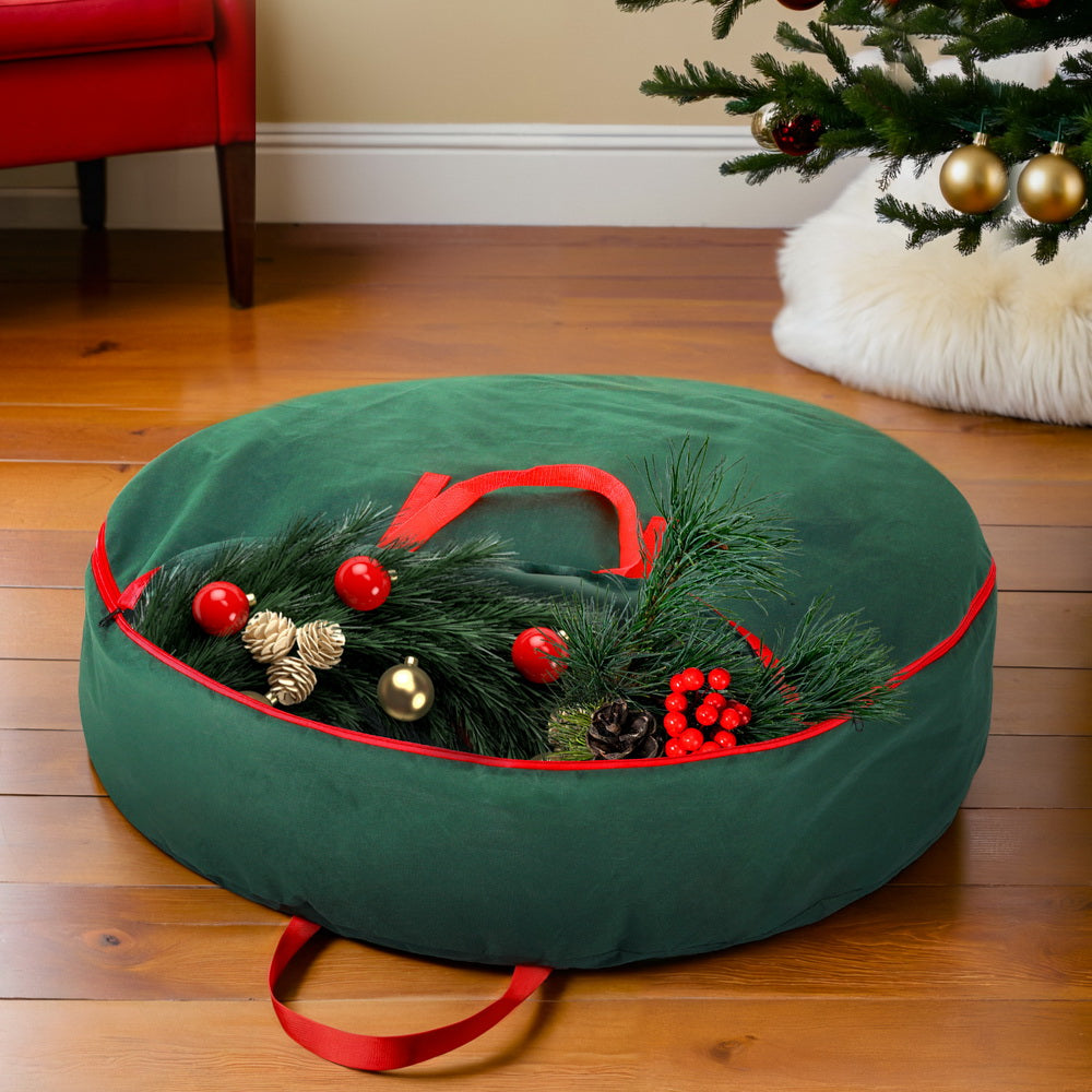 Christmas Garland Wreath Storage Bag Baubles Ornaments Container in green with red handles and zipper on wooden floor near Christmas tree