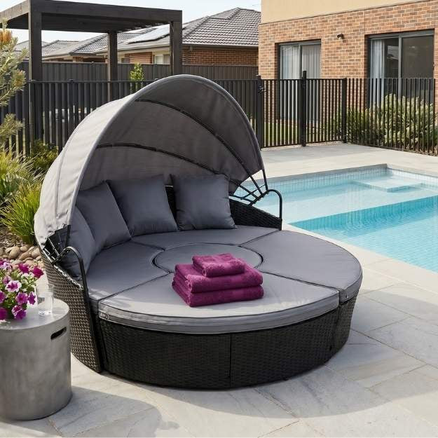 Bedzy outdoor wicker daybed with retractable canopy, grey cushions, and purple towels, placed on a paved patio next to a backyard swimming pool.