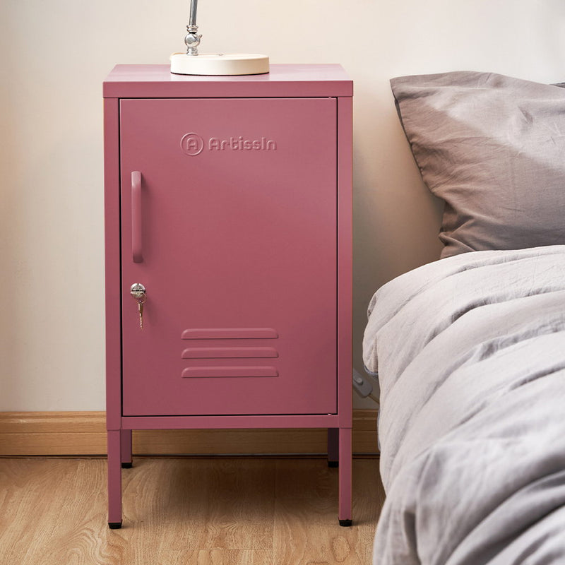 Bedside Table Metal Cabinet - MINI Pink with lockable door, metal construction, standing next to bed with gray bedding on wooden floor