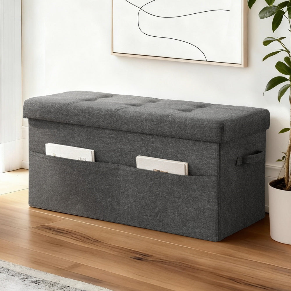 Storage Ottoman Foot Stool Rest Foldable in dark gray fabric with side pockets, tufted top, and handles in modern bedroom setting