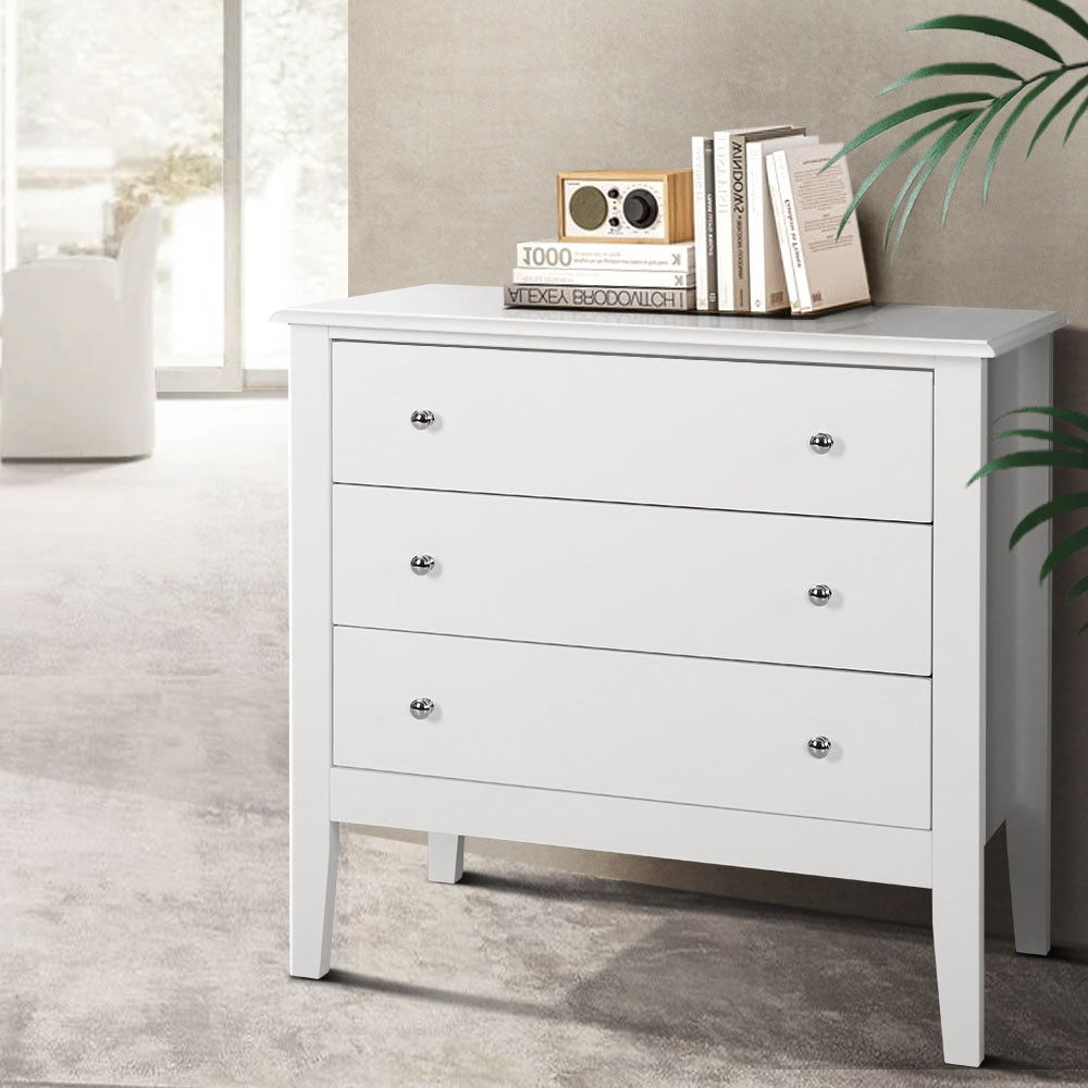Chest of 3 Drawers Storage Cabinet White with chrome knobs, standing on angled legs in a bright indoor bedroom setting