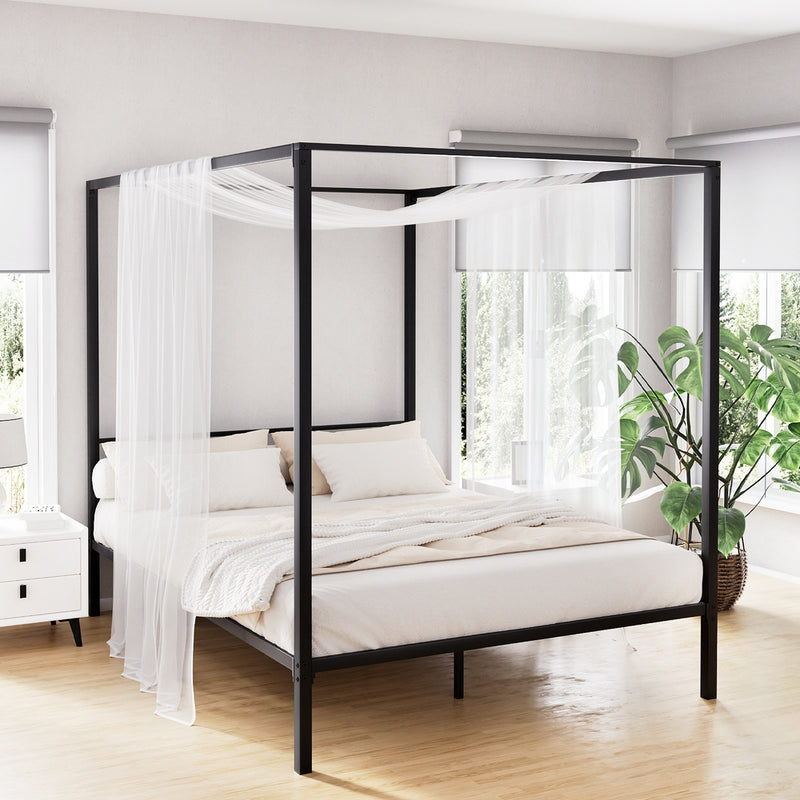 Metal Four Poster Double Size Bed Frame Black with sheer white curtains in bright bedroom featuring bedside table and plant