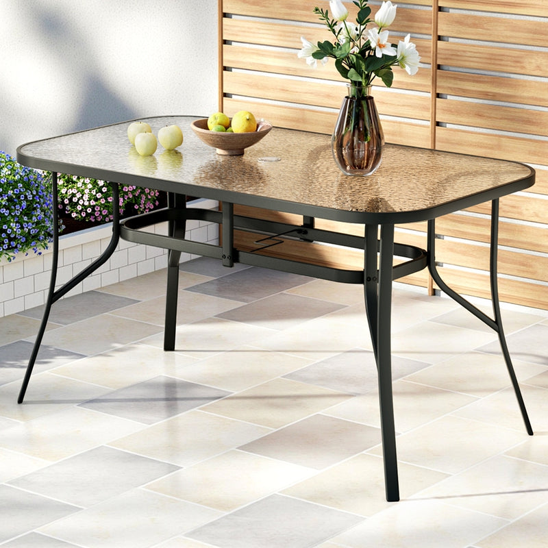 140cm Outdoor Dining Table With Glass Top and Parasol Hole – Black with black frame and decorative vase on tiled patio