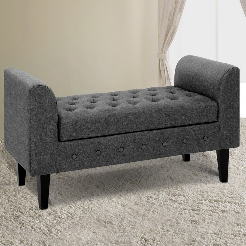 Fabric Storage Ottoman - Grey with tufted seat, button accents, and wooden legs, shown in a bedroom setting on carpet