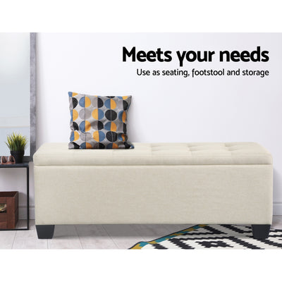 Large Fabric Storage Ottoman - Beige with tufted top, wooden feet, shown with patterned cushion and decorative rug in bedroom setting