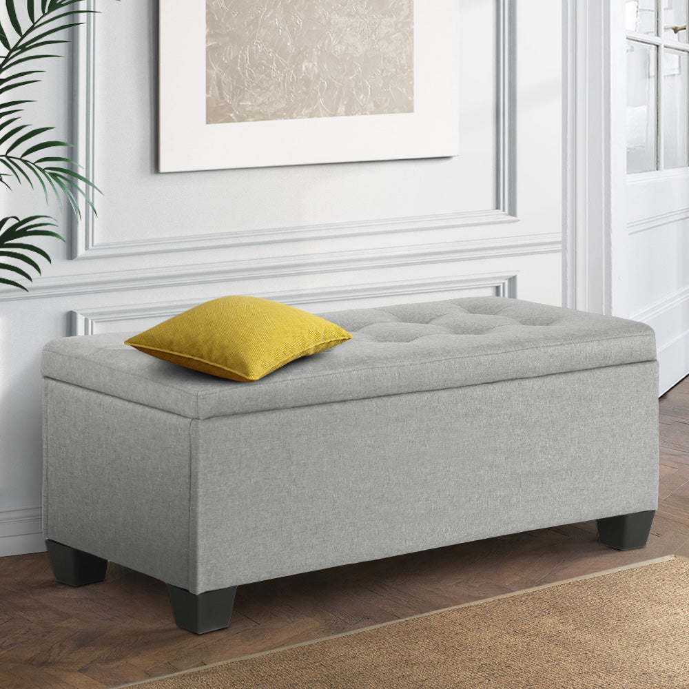 Large Fabric Storage Ottoman - Light Grey with tufted top and black feet, placed in a stylish bedroom setting with yellow cushion.