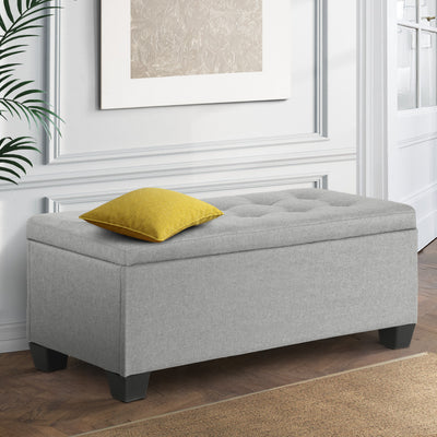Large Fabric Storage Ottoman - Light Grey with tufted top and black feet, placed in a stylish bedroom setting with yellow cushion.